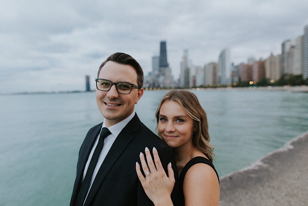 Photo of a couple standing at North Avenue Beach together and smiling at the camera while the woman shows off her engagement ring with Lake Michigan and the Chicago skyline in the background