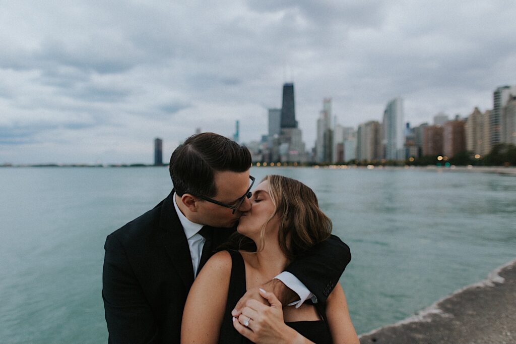 Photo of a couple kissing, the man is standing behind the woman as she holds his hand showing off her engagement ring, behind them is Lake Michigan and the Chicago skyline