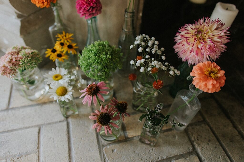 An assortment of different flowers sit in different shaped and sized glassware on a brick floor