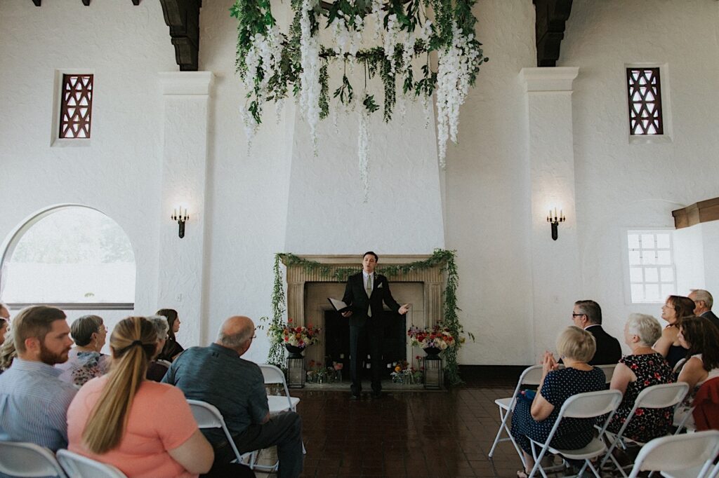A groomsmen who is also the officiant of the wedding speaks to seated guests in the indoor space of Venue 1929 in Springfield
