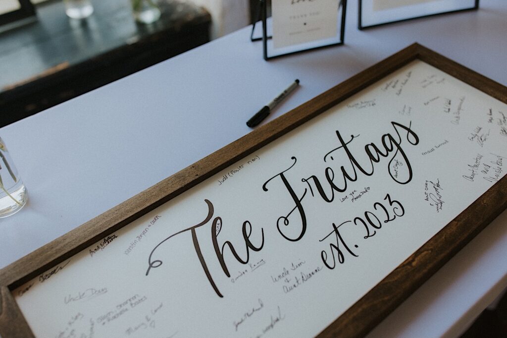 A sign in for wedding guests sits on a table, it is the couple's last name and their wedding date in a wood frame