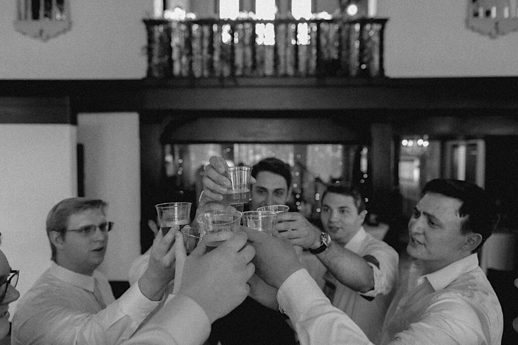 Black and white photo of a group of men raising plastic glasses together for a toast while inside Venue 1929 in Springfield during a wedding reception