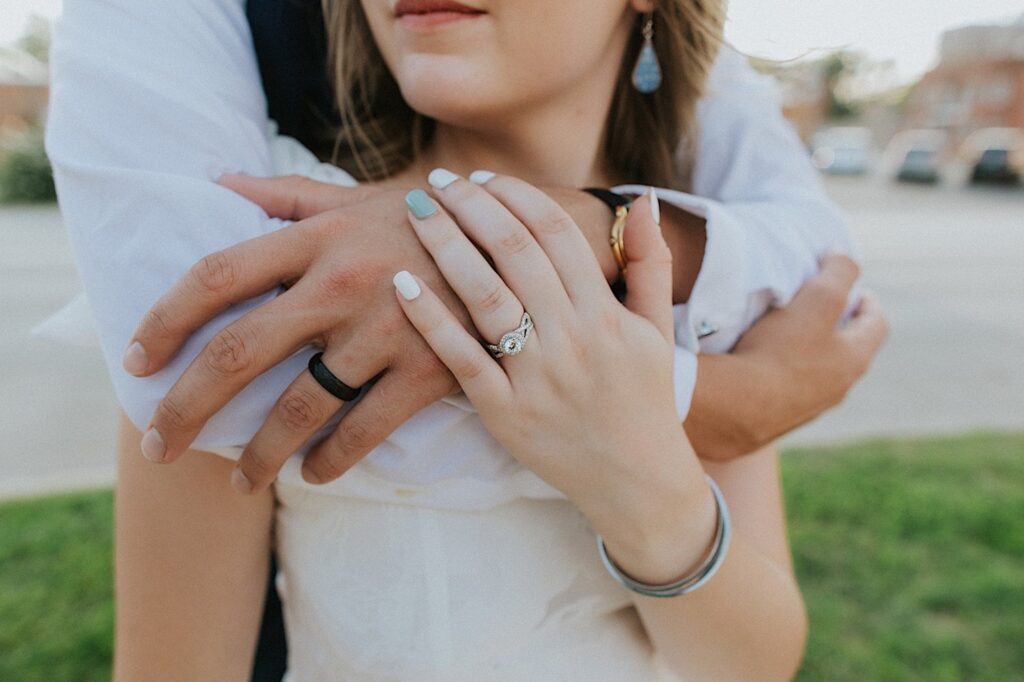 Close up photo of a bride and groom's hands resting on one another showing off their wedding rings as the groom hugs the bride from behind