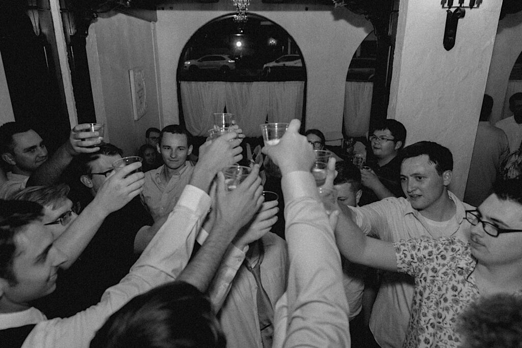 Black and white photo of guests of a wedding raising plastic glasses together for a toast during an indoor reception at Venue 1929 in Springfield