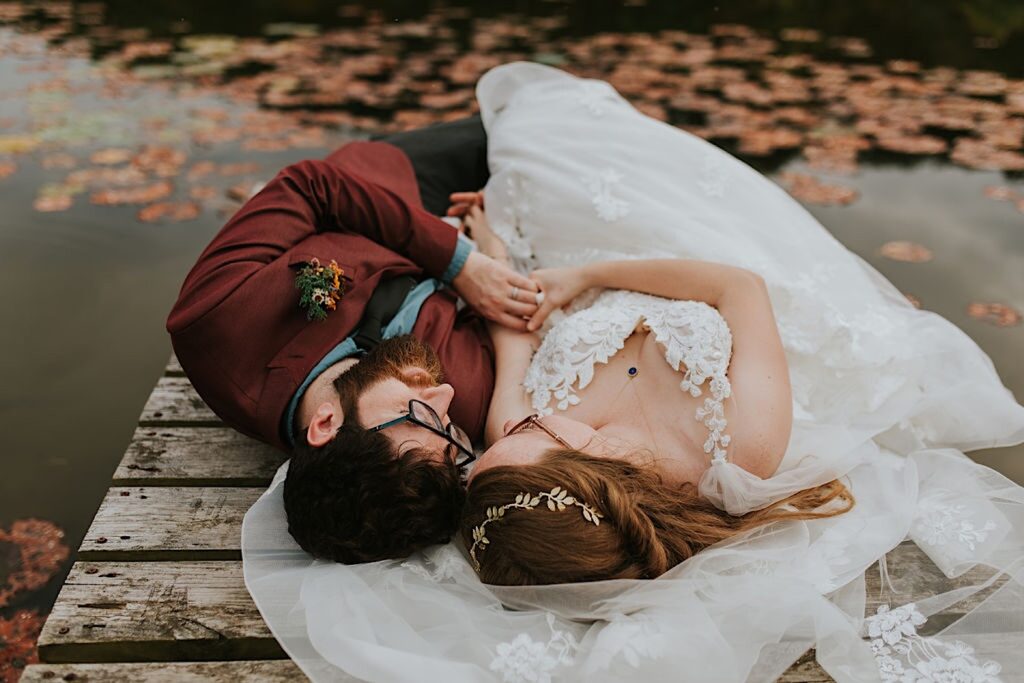A bride and groom lay together on a wooden dock next to a lake near their wedding venue out in nature