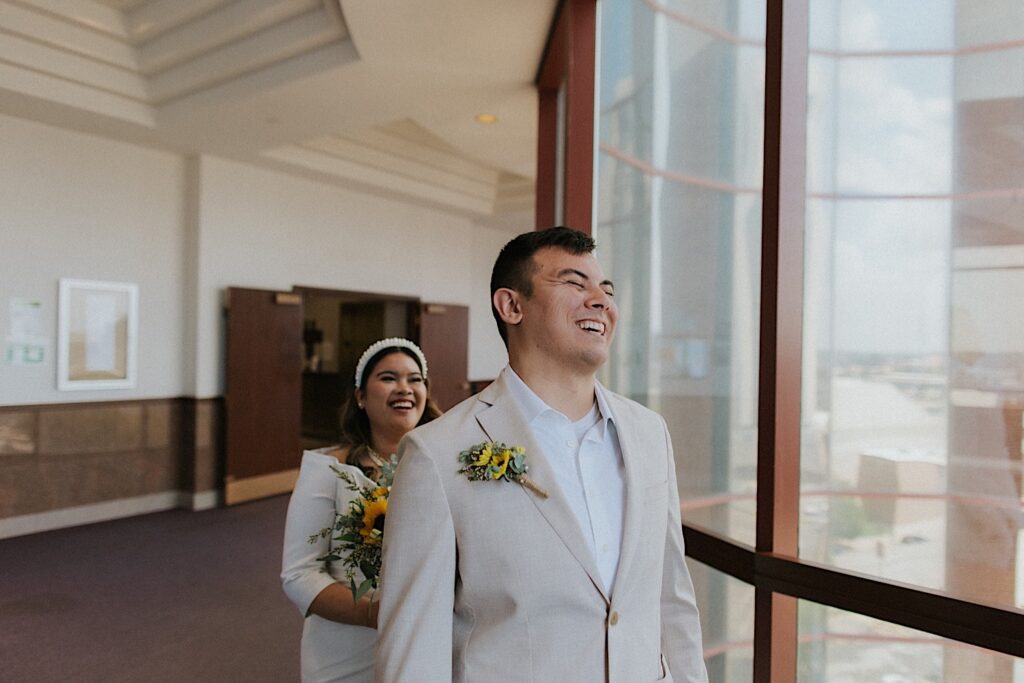 A groom laughs as the bride smiles behind him before their indoor first look for their wedding day at the Sangamon County Courthouse