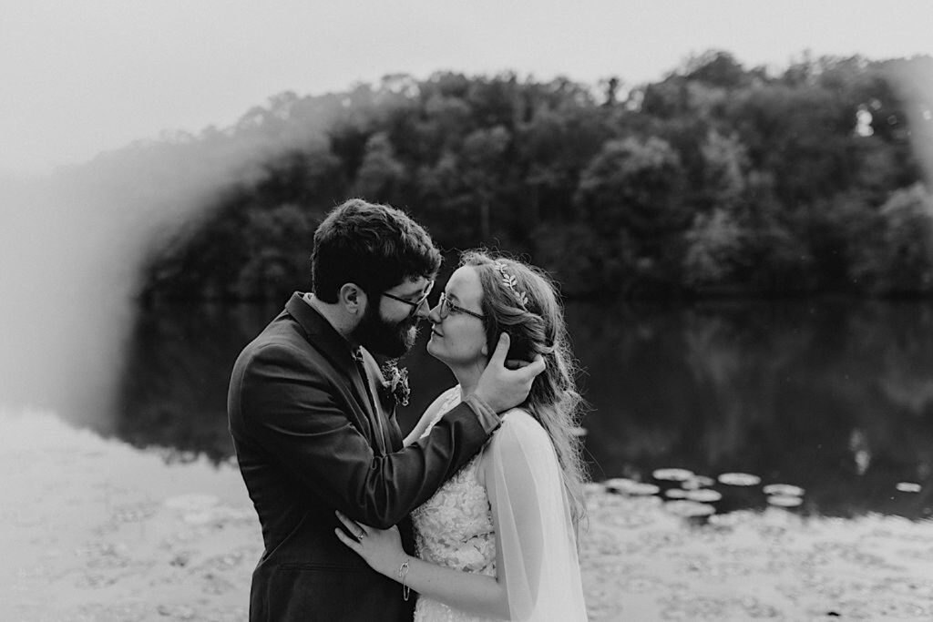 Black and white photo of a bride and groom about to kiss one another near their wedding venue which is on a lake surrounded by nature
