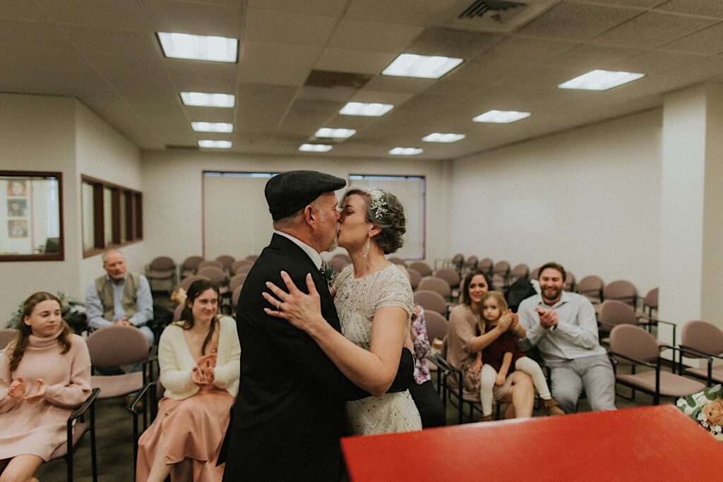 A man and woman kiss one another while in a courtroom for their elopement as guests seated in the background clap