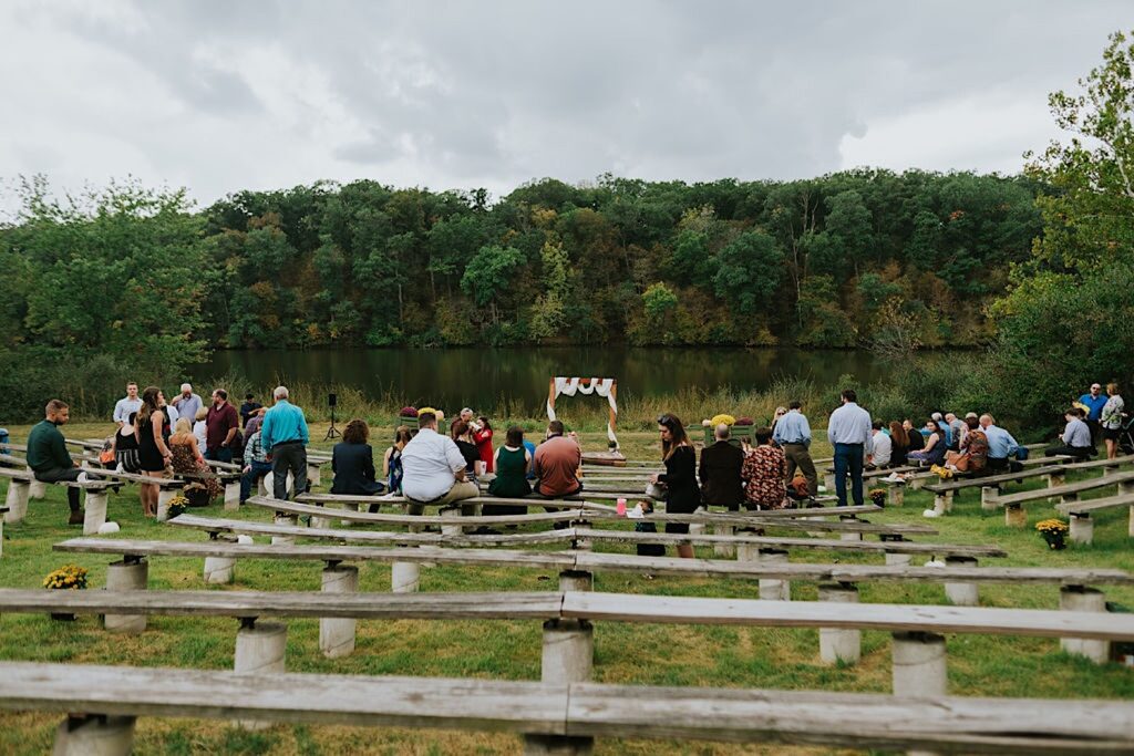 Guests of a wedding are seated at the ceremony space which looks out over a lake