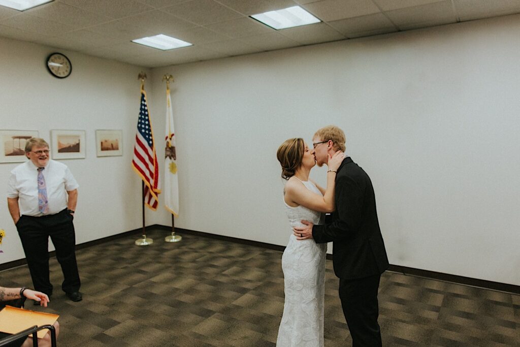 A bride and groom kiss one another while in a courtroom for their elopement as a guests watch from the side