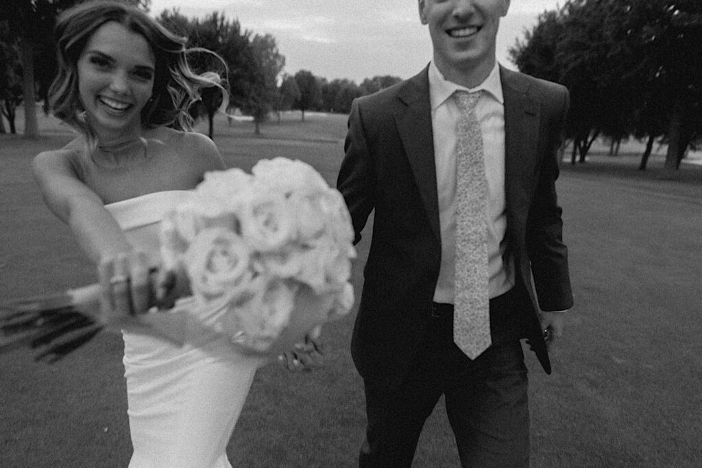 Black and white photo of a bride and groom smiling while on a golf course and walking towards the camera, the bride is extending her bouquet towards the camera