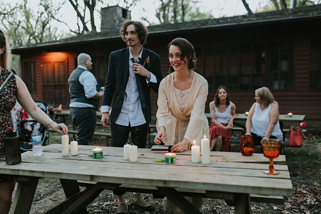 A bride and groom stand next to one another lighting candles at wooden tables at their outdoor wedding venue surrounded by nature