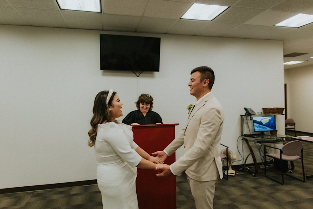A bride and groom smile at one another while holding hands during their elopement ceremony in a courtroom as their officiant smiles in the background