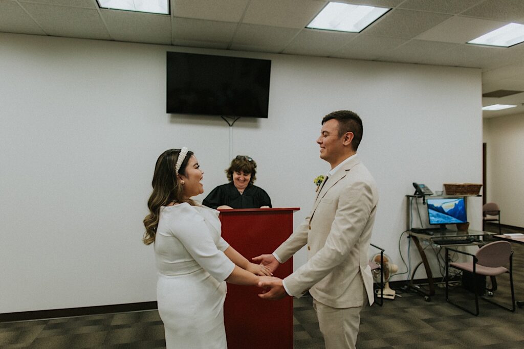 A bride and groom smile while holding hands and facing one another during their wedding ceremony at the Sangamon County Courthouse