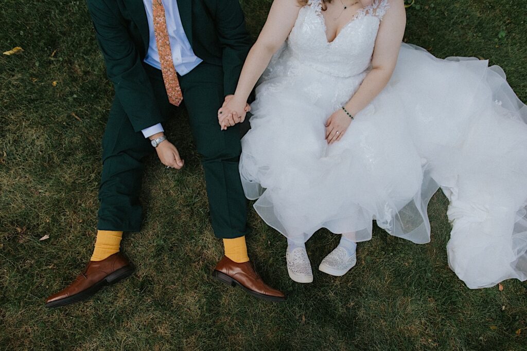 Top down photo of a bride and groom sitting next to one another in the grass and holding hands near their wedding venue, the Clayville Historic Site
