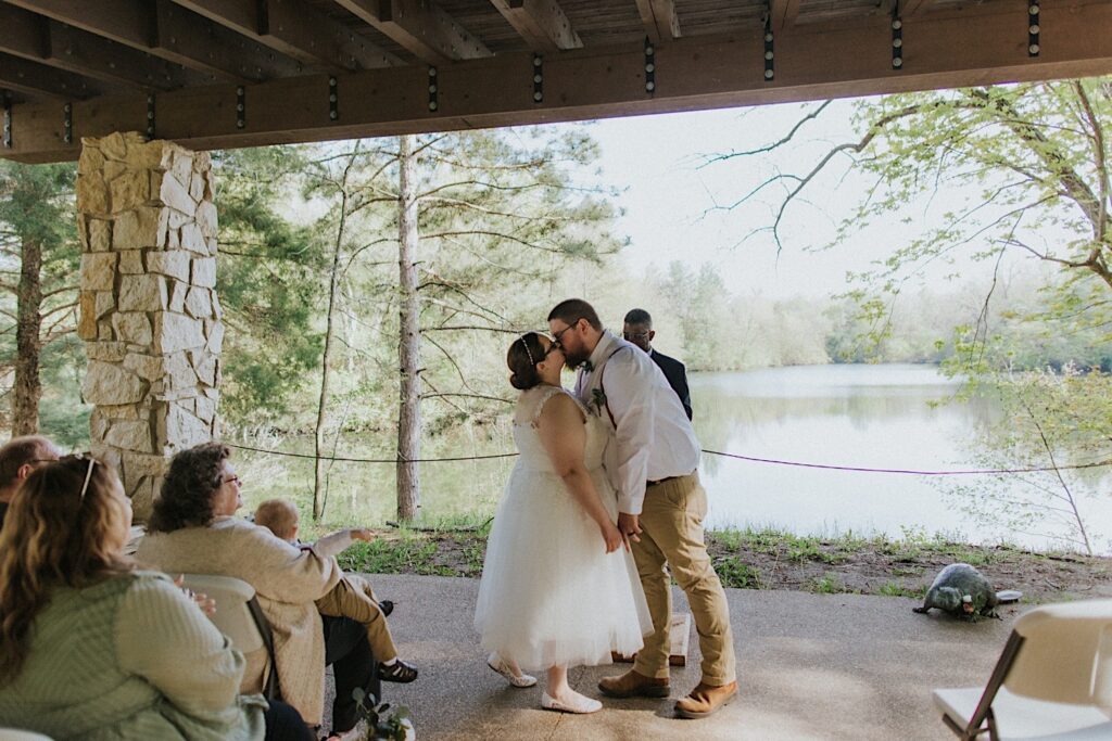 A bride and groom kiss one another during their wedding ceremony at their outdoor wedding venue looking out over a lake and surrounded by nature