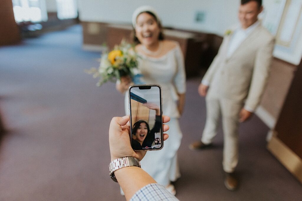 A woman who has is being facetimed on a phone waves while the bride and groom stand in the background
