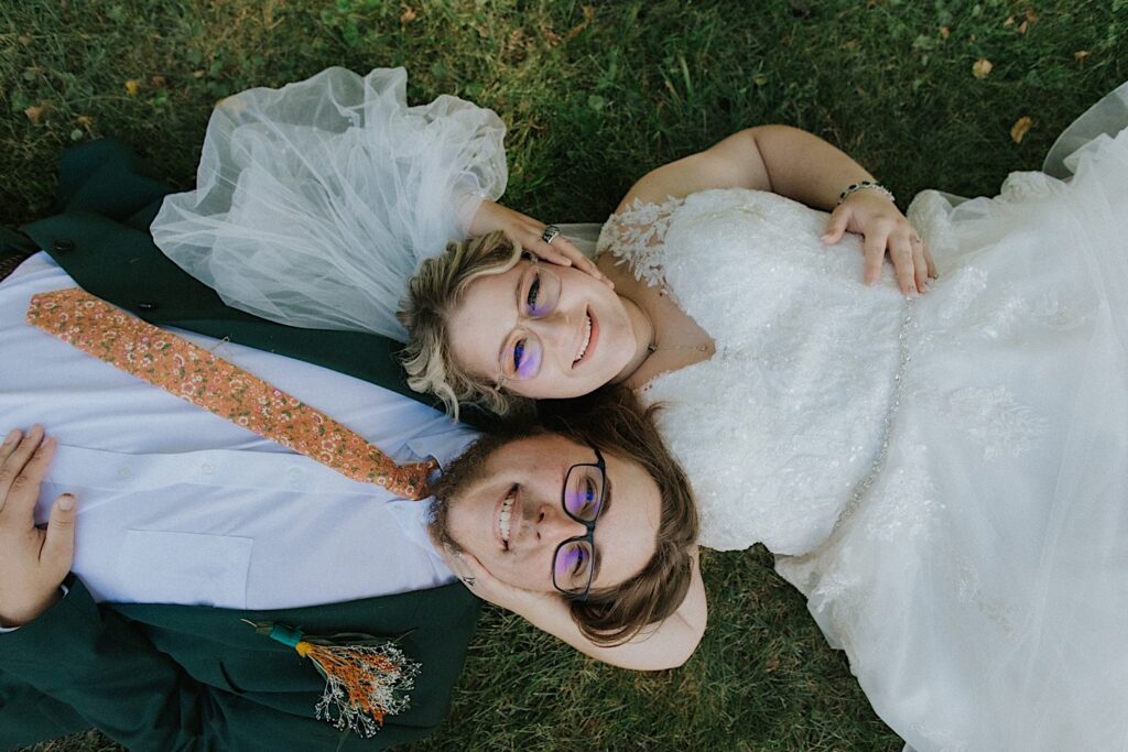 A bride and groom lay in the grass together with their heads next to one another as they smile up at the camera during their wedding day at the Clayville Historic Site