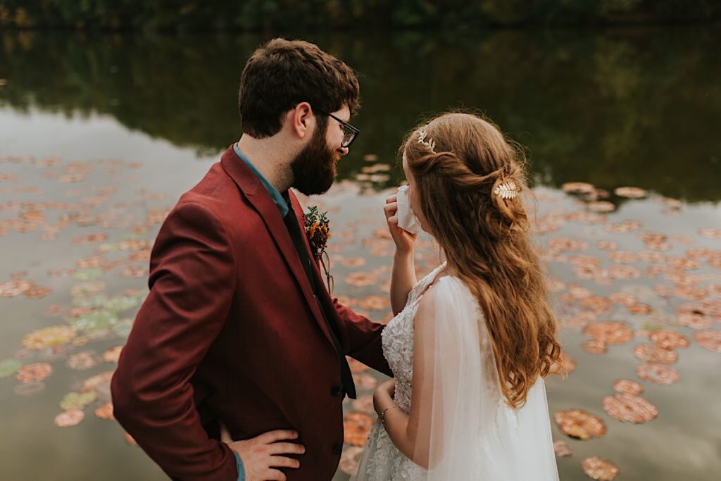 A bride wipes a tear from her eye as the groom comforts her while the two stand next to a lake