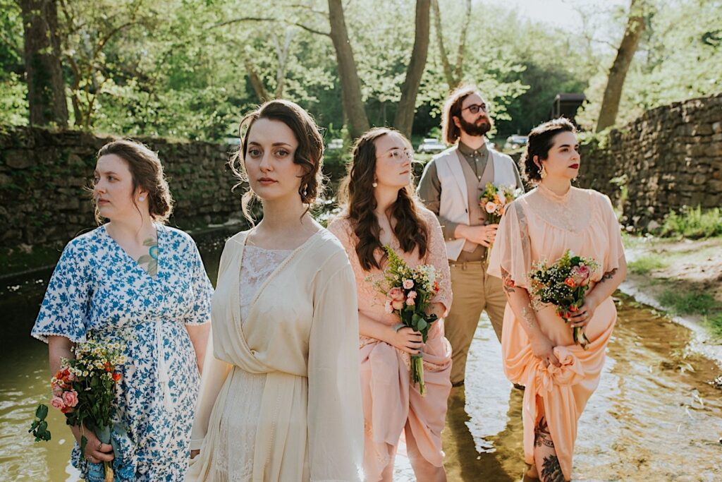 A bride stands with members of her wedding party posing dramatically while standing in a river