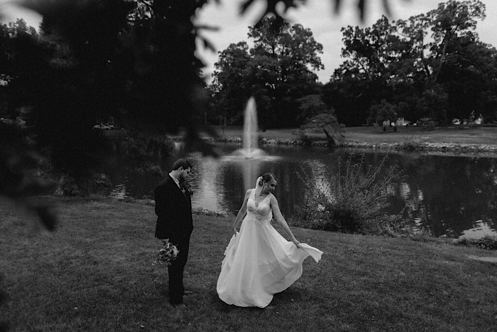 Black and white photo of a bride dancing in a field next to a pond with the groom standing at her side watching