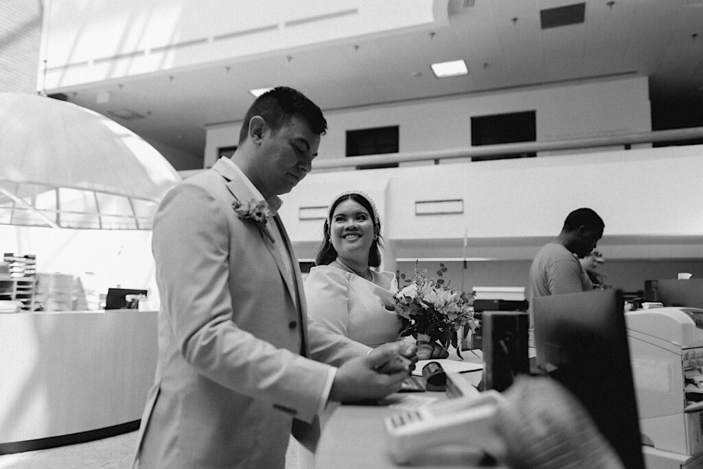 Black and white photo of a bride smiling at the groom while the two stand at a desk in the Sangamon County Courthouse getting their marriage license signed on their wedding day