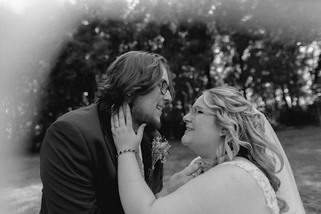 Black and white photo of a bride and groom smiling at one another while touching each other's faces to show off their wedding rings while outside their wedding venue, the Clayville Historic Site