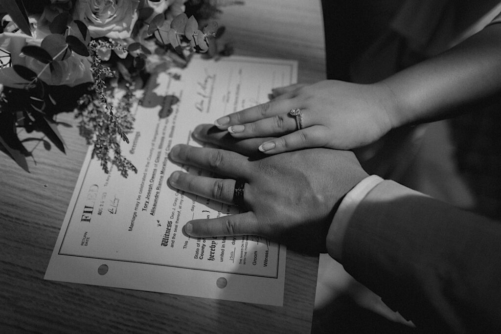 Black and white photo of a bride and groom putting their hands on their marriage license showing off their wedding rings after their intimate wedding at the Sangamon County Courthouse