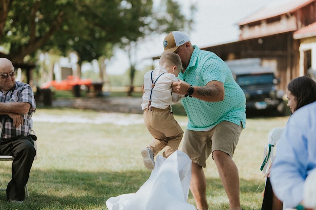 During an outdoor wedding ceremony at the Clayville Historic Site a man lifts a young boy in the air as a white cloth that is lining the aisle of the ceremony space sticks to the boy's shoe