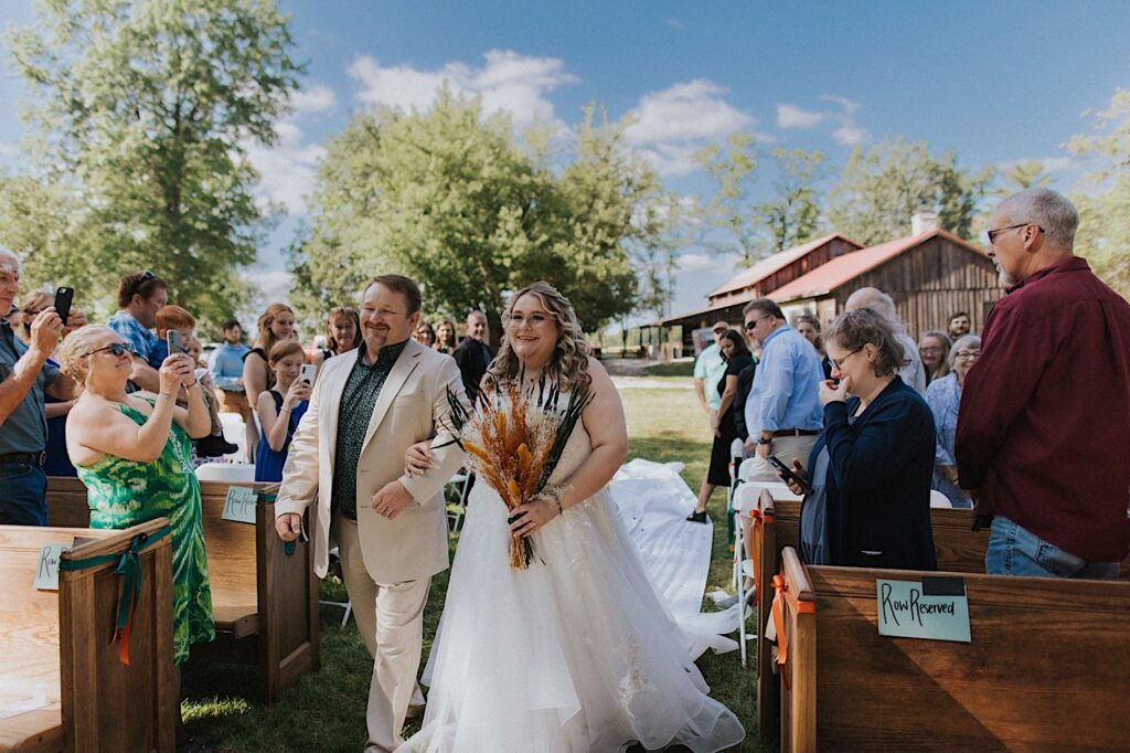 A bride smiles while walking down the aisle of her outdoor wedding ceremony at the Clayville Historic Site with her father as guests watch around her