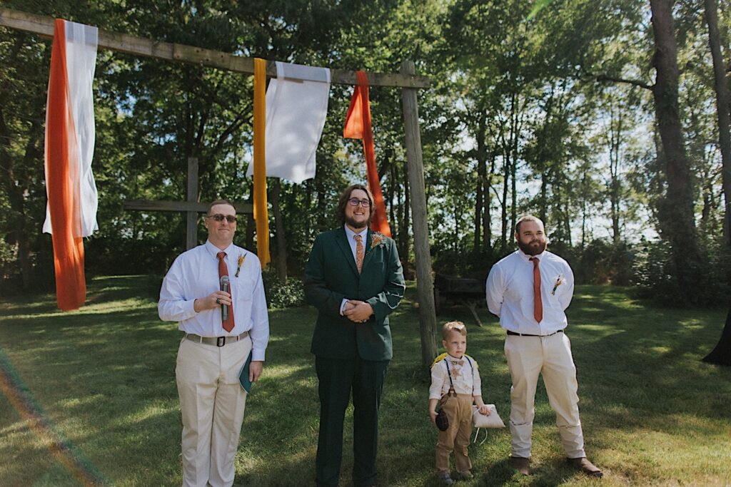 A groom smiles while standing at the outdoor ceremony space of his wedding at the Clayville Historic Site with his groomsmen and officiant on either side of him
