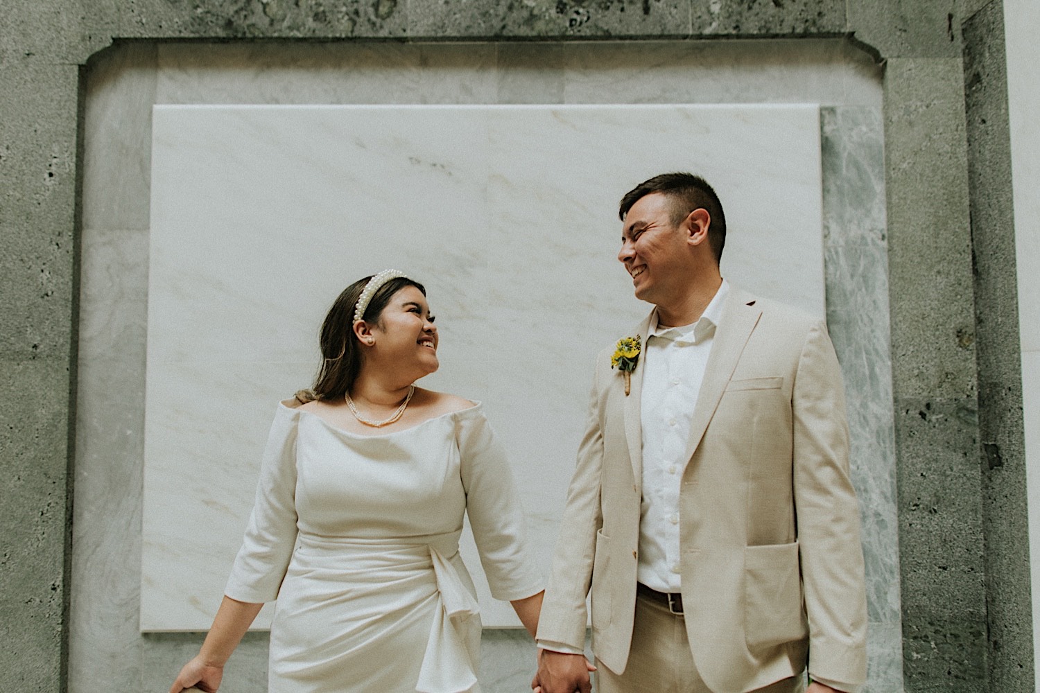 A bride and groom stand in front of a marble wall and smile at one another while inside the Illinois Capitol Building