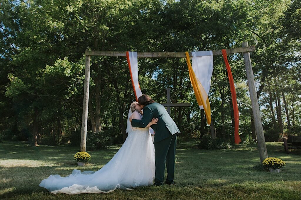A bride and groom kiss while underneath a wooden archway during their outdoor wedding ceremony at the Clayville Historic Site