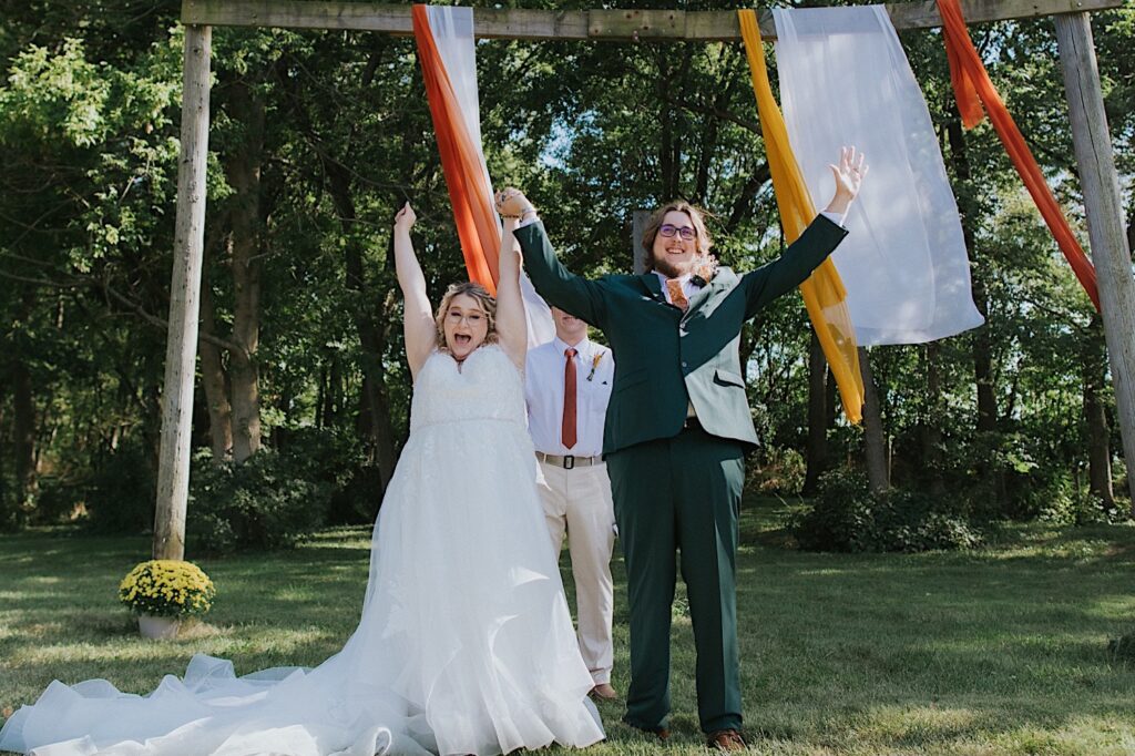 A bride and groom raise their arms and cheer in excitement after their outdoor wedding ceremony at the Clayville Historic Site