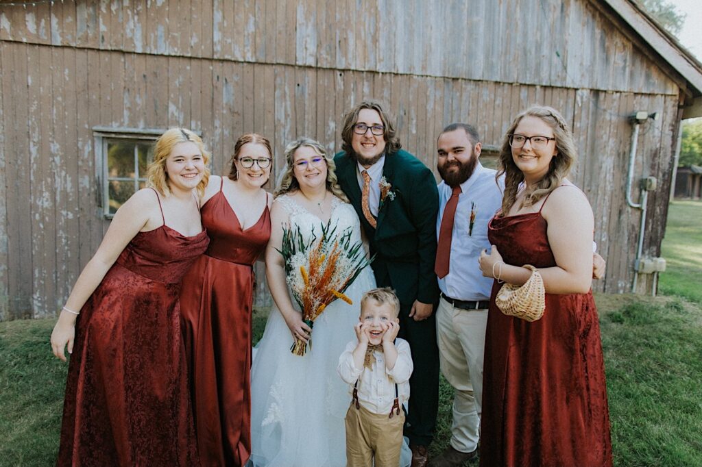 A bride and groom smile while surrounded by the members of their wedding parties as they stand outside a building of the Clayville Historic Site