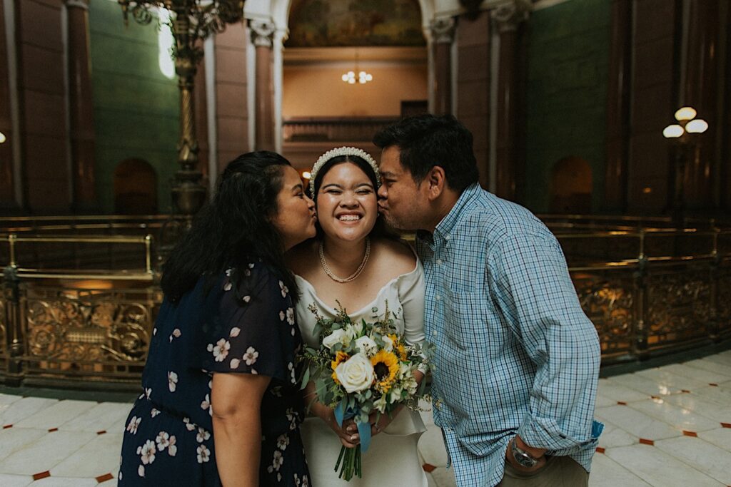 A bride smiles while inside the Illinois Capitol Building as her mother and father kiss her cheeks while standing on either side of her