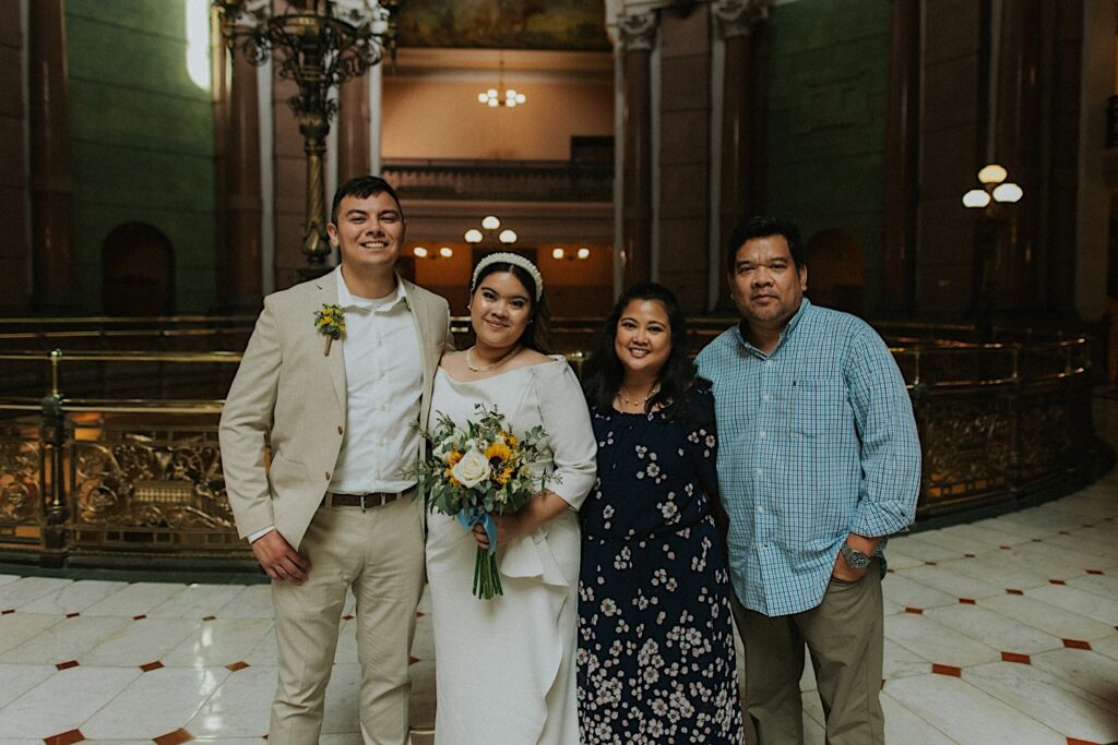 A bride and groom stand next to the bride's parents to pose for a photo while inside the Illinois Capitol Building