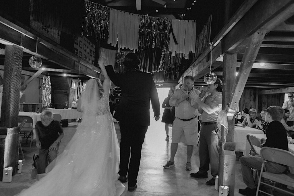 Black and white photo of a bride and groom entering their indoor wedding reception at the Clayville Historic Site as guests clap and cheer around them