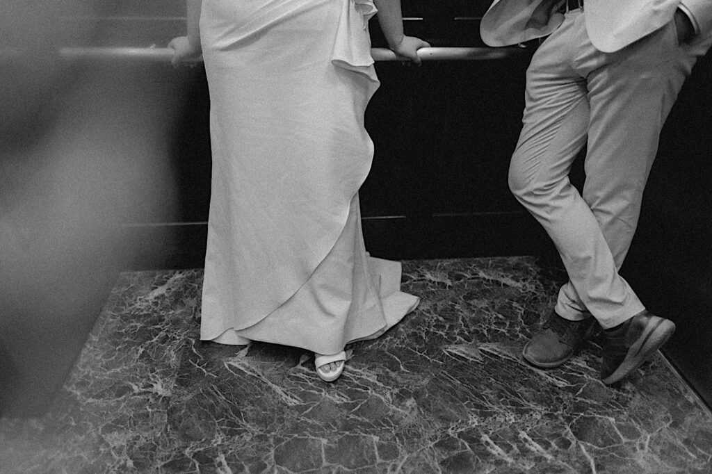 Black and white photo of a bride and groom's legs as they stand together in an elevator