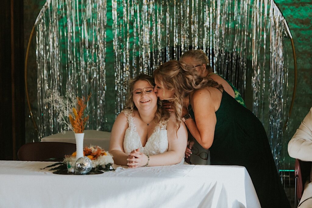 A bride smiles while getting kissed on the cheek by a woman while she sits at her sweetheart table during her indoor wedding reception at the Clayville Historic Site