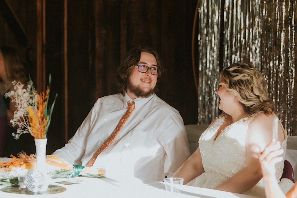 A bride and groom smile at one another while sitting at their sweetheart table during their indoor wedding reception at the Clayville Historic Site