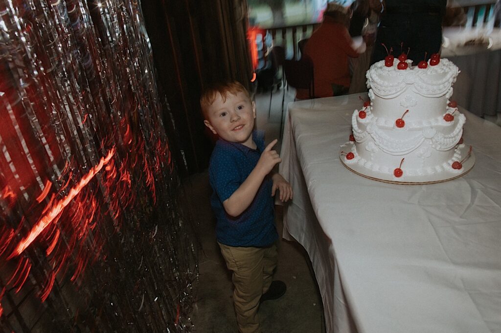 A young child smiles and points at a cake on a table during an indoor wedding reception at the Clayville Historic Site