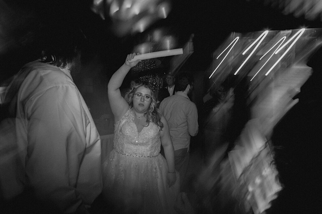 Black and white photo of a bride holding a glow stick in the air while dancing during her indoor wedding reception at the Clayville Historic Site