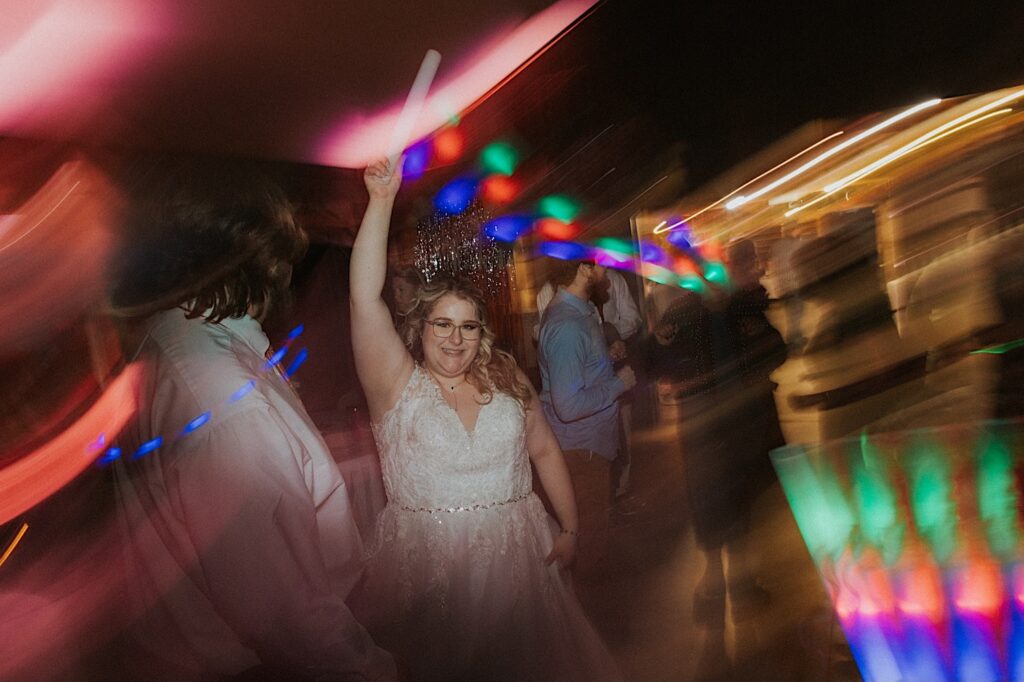 A bride smiles while holding a glowstick in the air while dancing during her indoor wedding reception at the Clayville Historic Site 