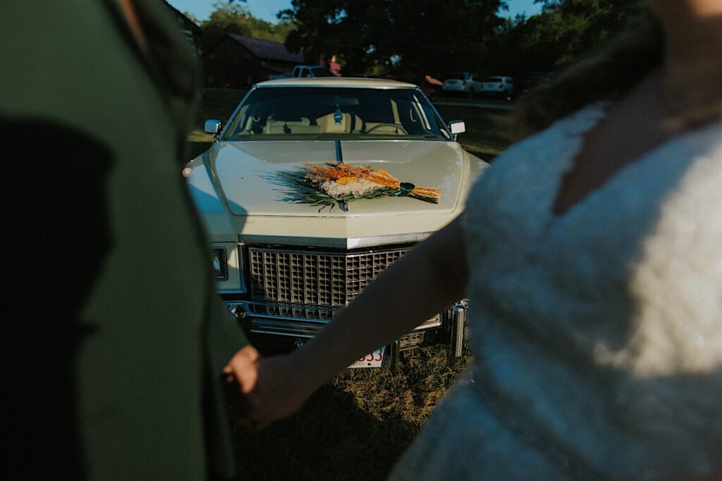 A bride and groom face one another in the foreground of the photo, behind them in focus is a classic car with their bouquet resting on the hood, photo taken at their wedding venue the Clayville Historic Site