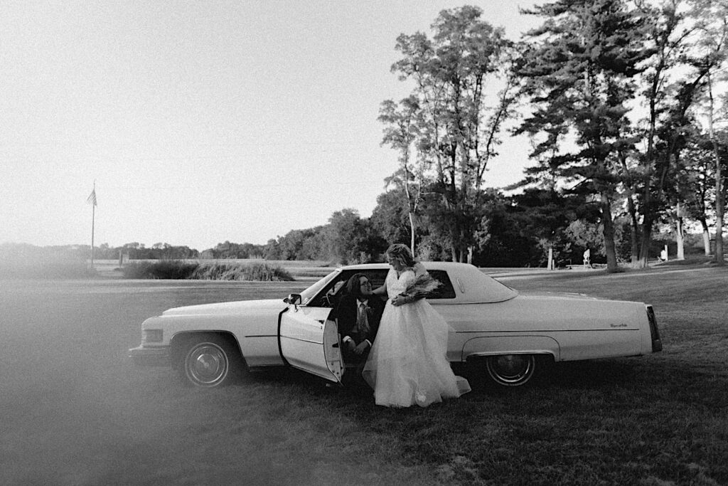 Black and white photo of a groom sitting in a classic car while the bride stands at his side during their wedding day at the Clayville Historic Site