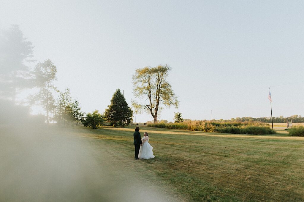 A bride and groom stand facing one another in a field at their wedding venue, the Clayville Historic Site while the sun sets behind them