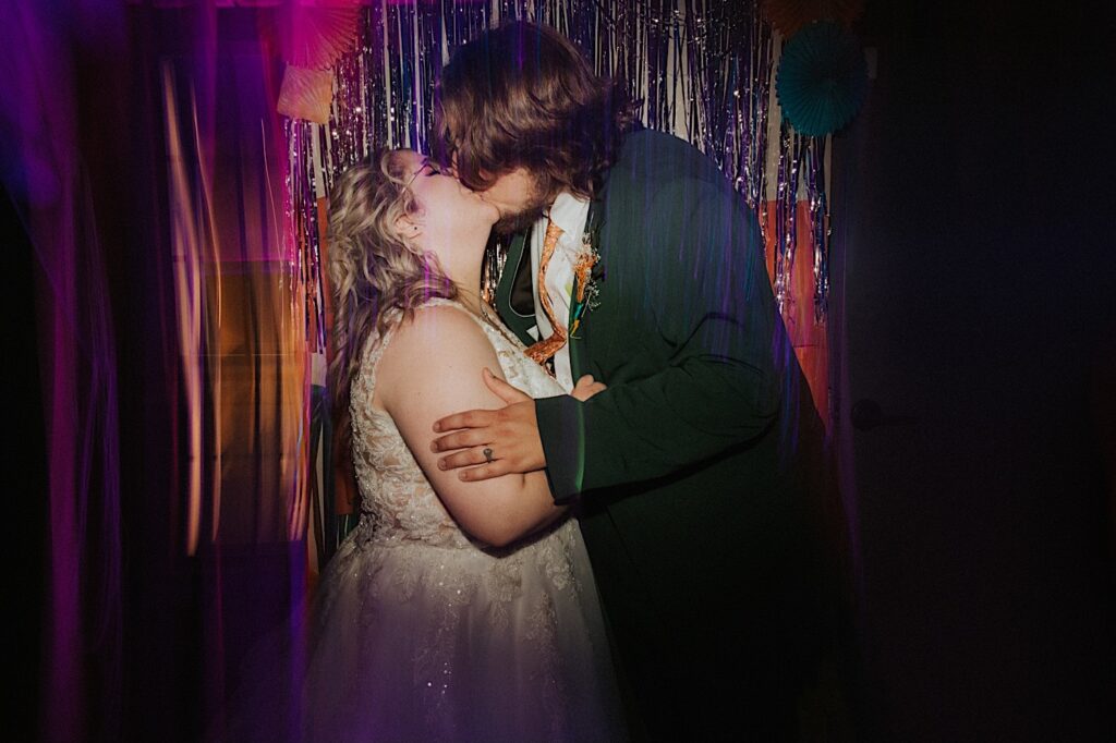 A bride and groom kiss one another while standing in front of a wall of glittery strands and colorful décor during their indoor wedding reception at the Clayville Historic Site