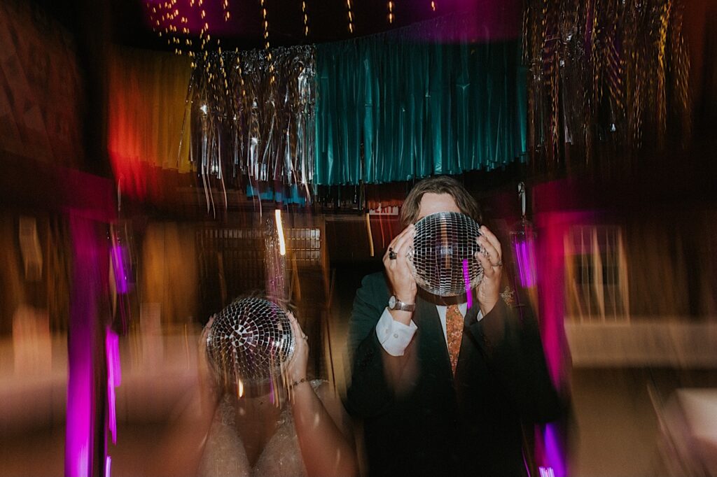A bride and groom hold disco balls in front of their faces while light distorts around them during their indoor wedding reception at the Clayville Historic Site