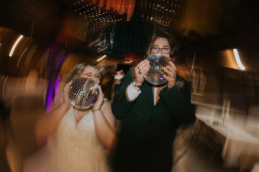 A bride and groom hold disco balls in front of their faces as they peek their eyes over while light distorts around them during their indoor wedding reception at the Clayville Historic Site